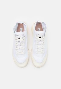 Vita idrottsskor med slät syntetisk ovandel, texturerade detaljer, rund logotyp på tungan och krämfärgade gummisulor. Snörena är platta och vita.