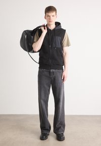 Μαύρο γιλέκο με φερμουάρ και γκρι τσέπη, φορεμένο πάνω από μπεζ μπλούζα. Συνδυασμένο με σκούρες γκρι παντελόνες φαρδιές και μαύρα παπούτσια, κρατώντας μια μαύρη τσάντα.