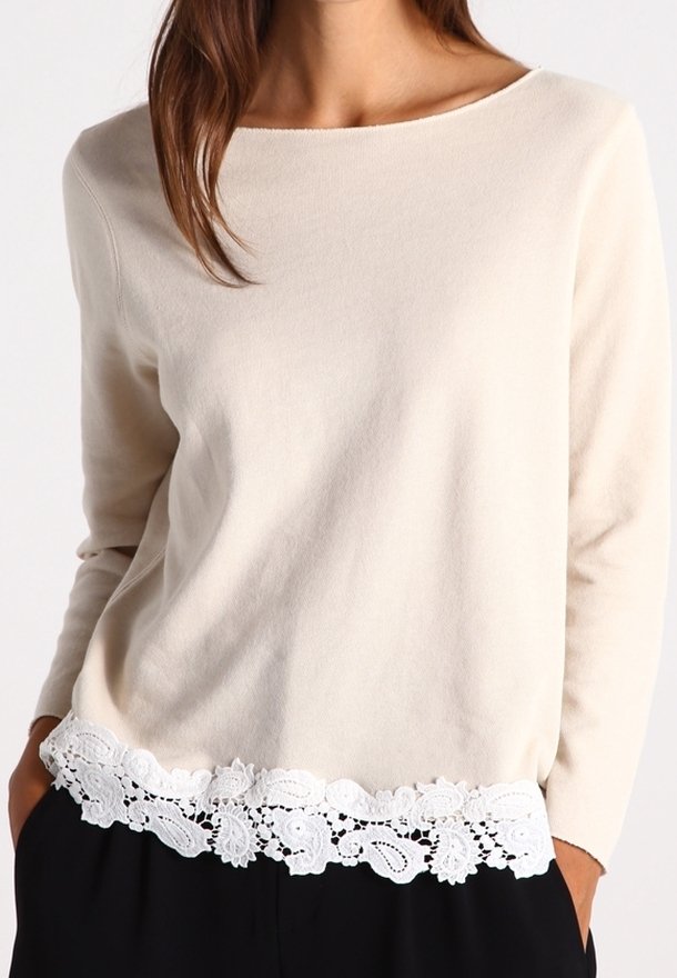 Femme portant un haut crème à manches longues avec bordure en dentelle blanche à l'ourlet, associé à un pantalon noir, mains dans les poches.