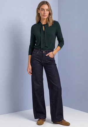 Une femme porte un chemisier vert foncé à nouer au cou, un jean foncé à jambes larges et des chaussures marron devant un fond de studio bleu pâle.