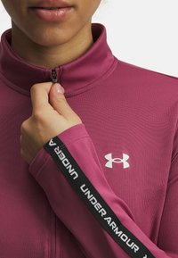 Fitted, zip-up athletic top in dark pink fabric with a mock neck, featuring white Under Armour logo on chest and woven black logo detailing on sleeves.