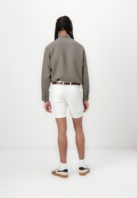 Vedere din spate a unei persoane îmbrăcate cu o cămașă gri cu mâneci lungi, pantaloni scurți albi și adidași albi cu accente negre; stând în fața unui fundal simplu.