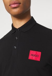 Gros plan sur une personne portant un polo noir avec une étiquette rectangulaire rouge portant le mot "HUGO" sur la poitrine.