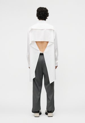 Person with curly hair wearing a white long-sleeve shirt with an open back over gray pants and black shoes with block heels.