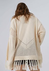 Beige Häkel-Cardigan mit offenem Strickmuster, versehen mit einem V-förmigen Design und Fransen am Saum. Lockerer Schnitt, lange Ärmel, strukturierter Stoff.