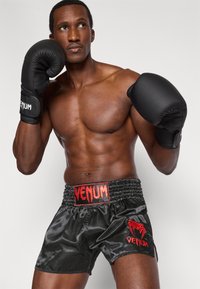 Fekete bokszkesztyűk és szatén fekete rövidnadrág piros VENUM logóval; a modell bokszállást vesz fel, bemutatva tónusos testalkatát és jól definiált vonalait.