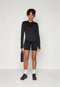 Schwarzes langärmliges Sportoberteil und Shorts-Set, kombiniert mit schwarzen Sneakers und einer schwarzen Wasserflasche. Merkmale sind Logodetails und ein figurbetontes Design.