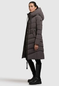 Lang puffet frakke i mørkegrå med horisontal quiltet design, oversized hætte, lynlåslommer og mærke hængende. Stylet med knæhøje sorte støvler.