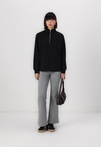 Svart zip-up hettegenser med lange ermer, kombinert med grå slengbukser. Modellen holder en mørkebrun skulderveske. Bakgrunnen er hvit.