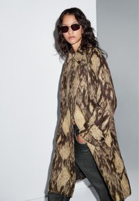 Brauner und beiger gemusterter oversized Mantel mit Knopf-Akzenten, kombiniert mit dunklen Jeans und dunklen rechteckigen Sonnenbrillen.