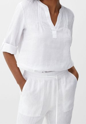 Vrouw draagt een wit linnen blouse met lange mouwen en opgerolde manchetten en bijpassende witte linnen broek met zakken en een trekkoord in de taille.