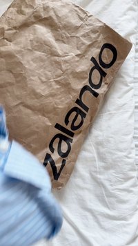 Barna, gyűrött papírcsomag, az oldalán merész fekete "zalando" felirattal, egy fehér, texturált felületen elhelyezve. Kék csíkos ing részben látható.