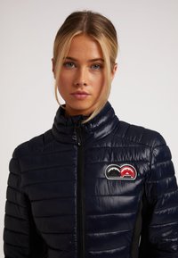 Marineblaue Daunenjacke mit horizontalen Nähten, hohem Kragen und Frontreißverschluss. Mit Logo-Patch in Rot und Schwarz auf der Brust.