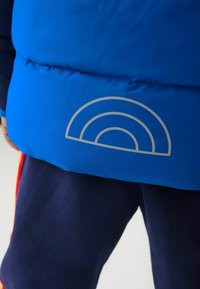 Fényes kék puffermellény sima textúrával, hátán ezüst szivárvány grafikával, sötét tengerészkék nadrág felett viselve.