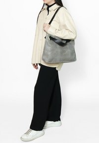 Borsa in pelle intrecciata grigia con una forma strutturata, dotata di una tracolla nera e una texture liscia. Indossata con una camicia crema e pantaloni neri.