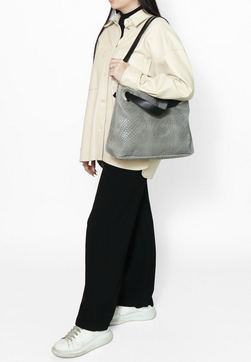 Borsa in pelle intrecciata grigia con una forma strutturata, dotata di una tracolla nera e una texture liscia. Indossata con una camicia crema e pantaloni neri.