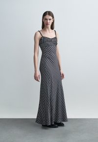 Žena v dlhej čiernej šatke bez rukávov s bielym geometrickým vzorom stojí na sivej podlahe pred jednoduchou svetlosivou stenou.