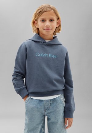Barn med blondt hår iført en blå Calvin Klein hættetrøje og lyseblå jeans, hænderne i lommerne, står mod en ensfarvet grå baggrund.