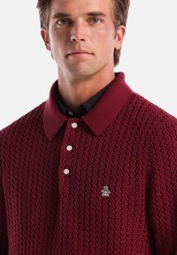 Burgunderfarbenes Poloshirt mit strukturiertem Strickmuster, weißen Knöpfen und einem Pinguin-Logo auf der Brust. Verfügt über einen gerippten Kragen und eine markante Form.