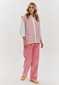 Różowa pikowana kamizelka z bocznymi kieszeniami, noszona na białej koszuli. Połączona z różowymi spodniami z otoman, a także żółtymi butami. Gładka faktura, swobodny styl.