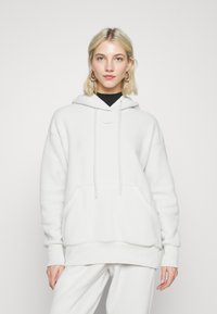 Vit fleece-hoodie med en framficka, snörning i huvan och en liten Nike-logga. Mjuk textur, avslappnad passform och ribbade ärmmuddar.