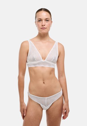 Femme à la peau claire et aux cheveux attachés porte un ensemble soutien-gorge en dentelle blanche et sous-vêtements assortis avec des motifs géométriques, debout devant un fond clair uni.