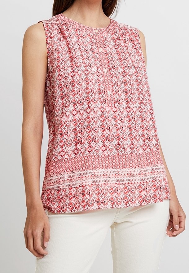 Blouse sans manches rouge et blanche à motif, avec un col rond et une patte de boutonnage, portée avec un pantalon blanc sur une personne debout.