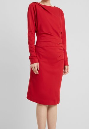 Vrouw draagt een rode jurk met lange mouwen tot op de knie met een licht geplooid detail in de taille tegen een effen grijze achtergrond.