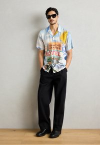 Krátkohlačasta, večbarvasta srajca s gumbi z vintage grafiko motela na svetlo modri tkanini, v kombinaciji s črnimi širokimi hlačami in črnimi čevlji.