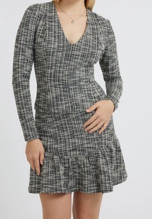 Kobieta ubrana w czarno-białą, kraciastą sukienkę z długim rękawem, głębokim dekoltem w szpic i falbaną u dołu, ozdobioną naszyjnikiem z wisiorkiem w kształcie serca.