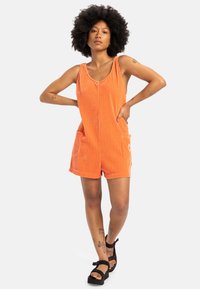 Oranje corduroy rompertje met dunne bandjes, twee voorzakken en een relaxte pasvorm. Gepaard met zwarte platform sandalen.