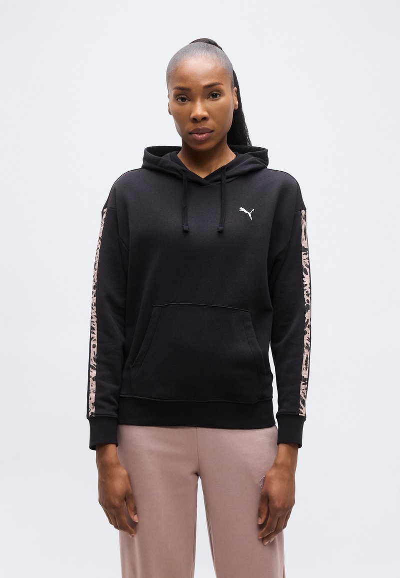 Γυναίκα που φοράει μαύρο hoodie Puma με ρίγες animal print στα μανίκια και ανοιχτό καφέ παντελόνι, στέκεται μπροστά σε απλό φωτεινό φόντο.