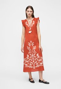 Γυναίκα που φοράει φορέμα μεσαίου μήκους χωρίς μανίκια σε σκουρόχρωμο πορτοκαλί με λευκή κεντημένη λουλουδάτη διακόσμηση, λαιμόκοψη με φούντες και μαύρα επίπεδα παπούτσια, στέκεται μπροστά σε απλό φόντο.
