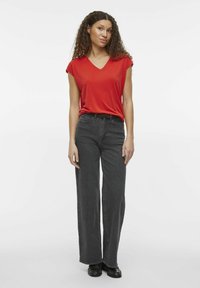 Camiseta roja de cuello en V combinada con unos pantalones oscuros de tiro alto y pierna ancha en gris oscuro. La modelo está de pie de frente sobre un fondo blanco liso.