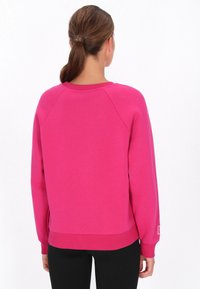 Sudadera fucsia con mangas raglán, cuello redondo y dobladillo acanalado. Cuenta con un pequeño parche de logo en la manga izquierda. Textura suave.