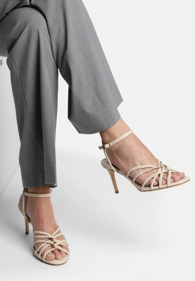 Sandales à talons hauts beiges avec des lanières entrelacées, bout ouvert et strap au niveau de la cheville. Le talon est fin et élégant, mettant en valeur un design classique.