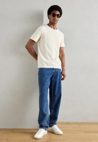 Tricou bej deschis cu mânecă scurtă, blugi bleu, largi, și adidași albi. Persoana poartă ochelari de soare rotunzi și stă în fața unui fundal neutru.