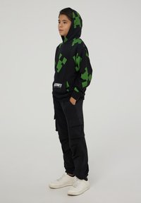 Felpa nera con cappuccio e motivo pixelato verde, tasca a marsupio, indossata con pantaloni cargo neri larghi e sneaker bianche.