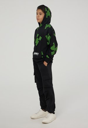 Felpa nera con cappuccio e motivo pixelato verde, tasca a marsupio, indossata con pantaloni cargo neri larghi e sneaker bianche.