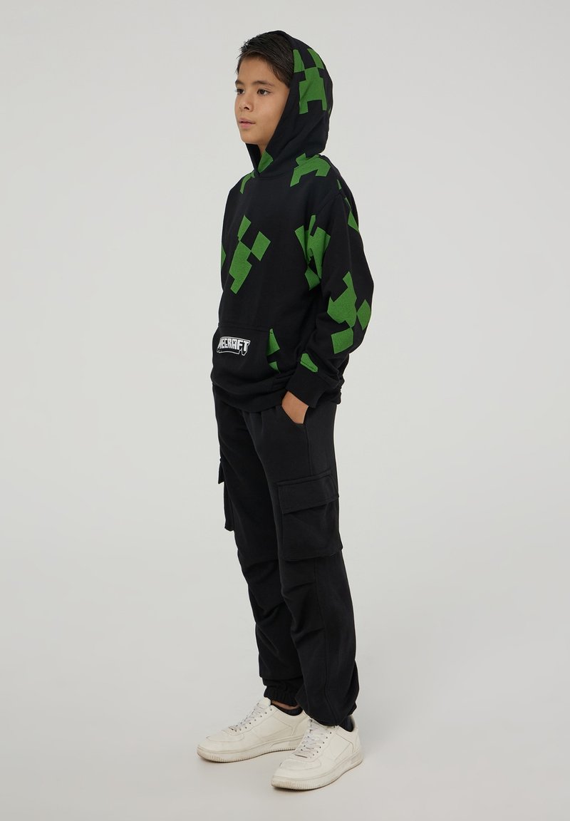 Felpa nera con cappuccio e motivo pixelato verde, tasca a marsupio, indossata con pantaloni cargo neri larghi e sneaker bianche.