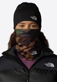 Beanie nera e gaiter per il collo mimetico con motivi marroni, verdi e neri, indossato con una giacca nera con zip e un top viola a trama.