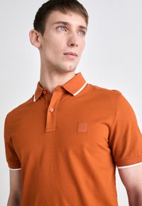 Oranžinė polo marškinė su struktūrizuota apykakle, dviem mygtukais ir balta juostele apykakle ir rankovėmis. Turi kvadratinį logotipo pleistrą.