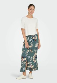 Ung kvinde med samlet hår iført hvid kortærmet skjorte, blomsteret knap-ned midi-nederdel, hvide sneakers og gyldne hoops-øreringe, stående.