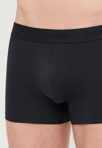 Schwarze Bio-Baumwoll-Boxershorts mit enganliegendem Design, ausgestattet mit einem elastischen Taillenbund mit "Organic Basics"-Markenlogo. Glatte Textur und keine Muster.