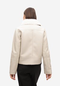 Giacca in finta pelle color crema con colletto in pelliccia sintetica, taglio cropped e dettagli di cucitura visibili. Indossata su pantaloni neri texture, vista da dietro.