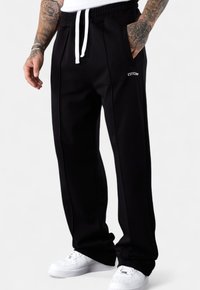 Man wearing black joggers with white drawstrings, white sneakers, and tattooed arms against a plain white background.