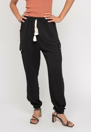 Femme portant un pantalon jogger cargo noir avec des chevilles élastiques et un cordon de serrage, assorti à des sandales à talons hauts noires à lanières et à un haut couleur rouille.