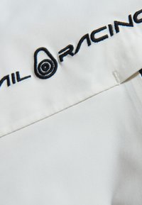 Vit tyg med svart broderad text och logotyp med cirkulärt detalj. Slät textur med noggrant sydda detaljer. Inga ytterligare mönster.