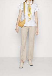 Mujer con camisa blanca de manga corta, pantalones beige con aberturas frontales, zapatos de lona beige, bufanda amarilla y bolso amarillo frente a un fondo blanco.