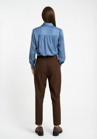 Niebieska satynowa bluzka z długimi rękawami i prostym krojem, zestawiona z brązowymi spodniami o dopasowanym kroju i ciemnymi butami, widziana z tyłu.